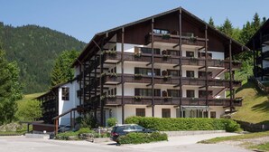 A multi-story hotel with balconies, a parking area, and a mountainous backdrop.