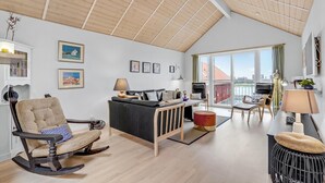 A modern living room with a sloped ceiling, a sofa, a rocking chair, a dining table, and a view of the outside.