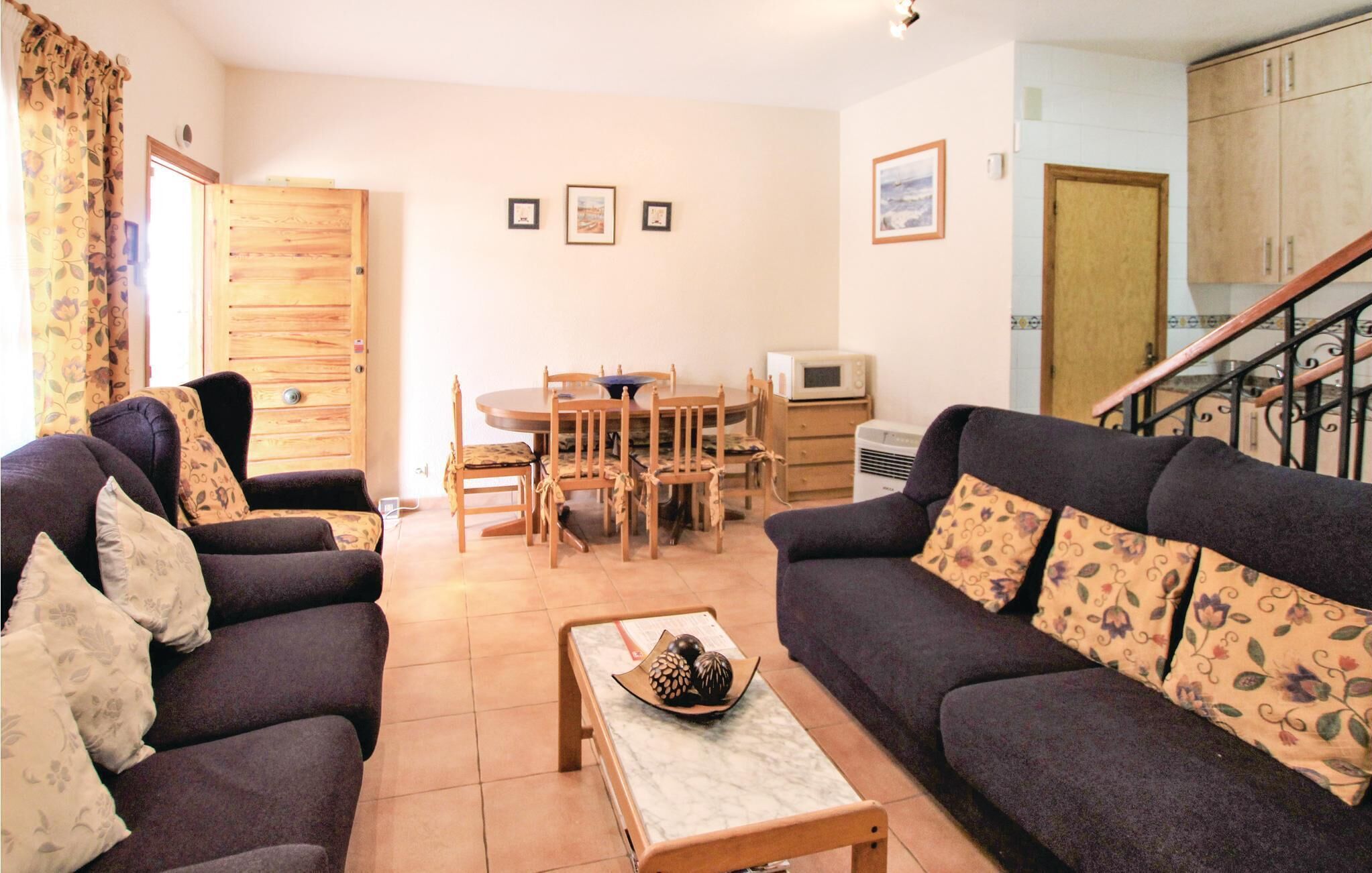 A living room with a sofa set, a dining table with chairs, a kitchen area with a microwave, and a staircase.