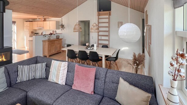 Un salon moderne avec un canapé d’angle gris, un coin repas avec une table blanche et des chaises noires, et une cuisine en arrière-plan.