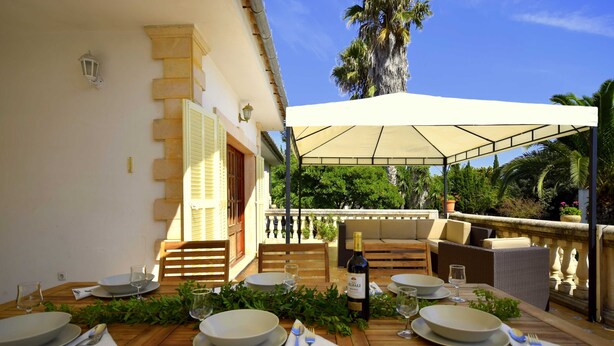 Un espace de restauration en plein air avec une table dressée pour quatre personnes, une bouteille de vin et un auvent offrant de l’ombre.