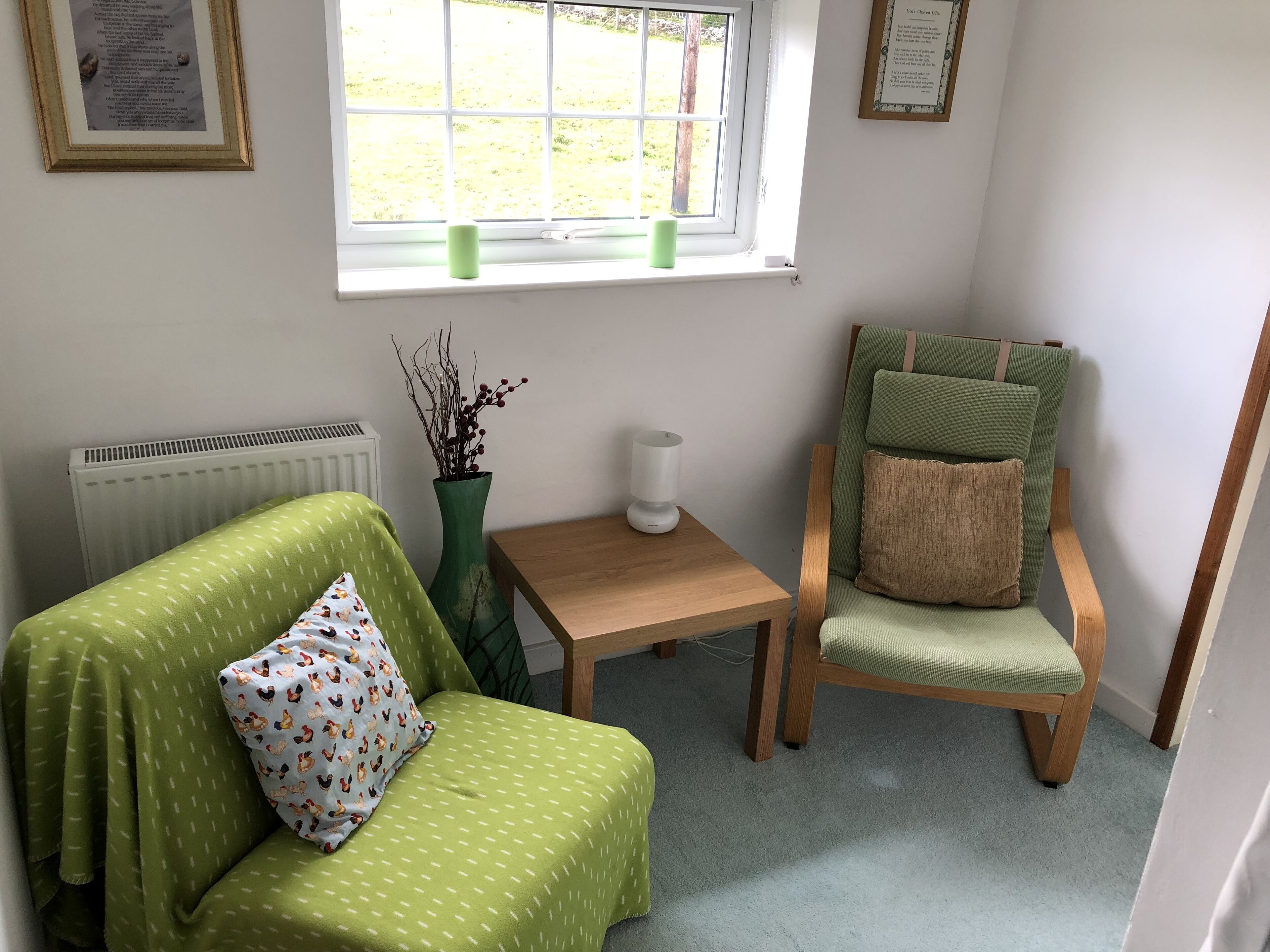 A small, cozy room with a green sofa, a wooden chair, a small table, a radiator, and a window with a view of a garden.