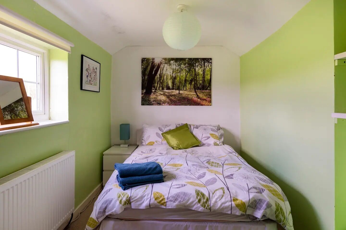 A bedroom with a bed, a nightstand, a radiator, a window, and a wall-mounted picture.