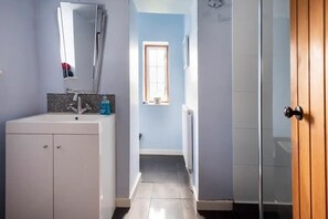 Ein Badezimmer mit weißem Waschbecken, Spiegel und Fliesenwand. Ein Fenster bietet Blick ins Freie, und eine Holztür befindet sich rechts.