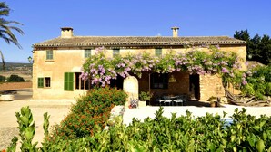 A stone building with a tiled roof, green shutters, and a garden with flowering plants. - 763424 Finca Conies - 763424 Ses Conies (Manacor)