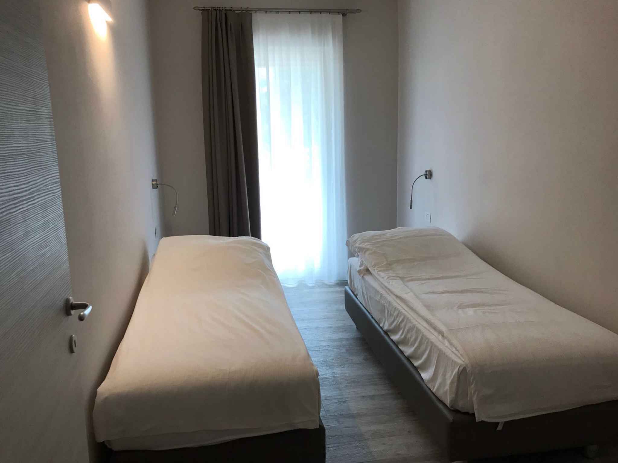 A small, single-room hotel room with two single beds, a window with curtains, and a door with a handle.