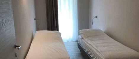 A small, single-room hotel room with two single beds, a window with curtains, and a door with a handle.