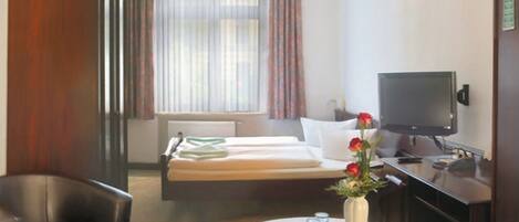 A hotel room with a bed, a desk with a television, a chair, a table with a vase, and a window with curtains.