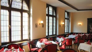 Un comedor con mesas dispuestas para el servicio, sillas tapizadas rojas y ventanas amplias con vidrieras decorativas.