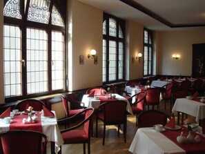 A dining area with tables set for service, red upholstered chairs, and large windows with decorative glass panes.