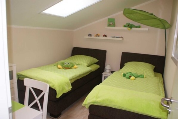 Une petite pièce bien éclairée, avec deux lits, chacun agrémenté de draps verts et d’un coussin à motifs de grenouilles. On y trouve une chaise blanche, une petite table et une étagère remplie de jouets.