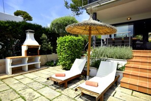 Een terras met twee loungestoelen, een rieten parasol, een schoorsteen en een eethoek met tafels en stoelen.