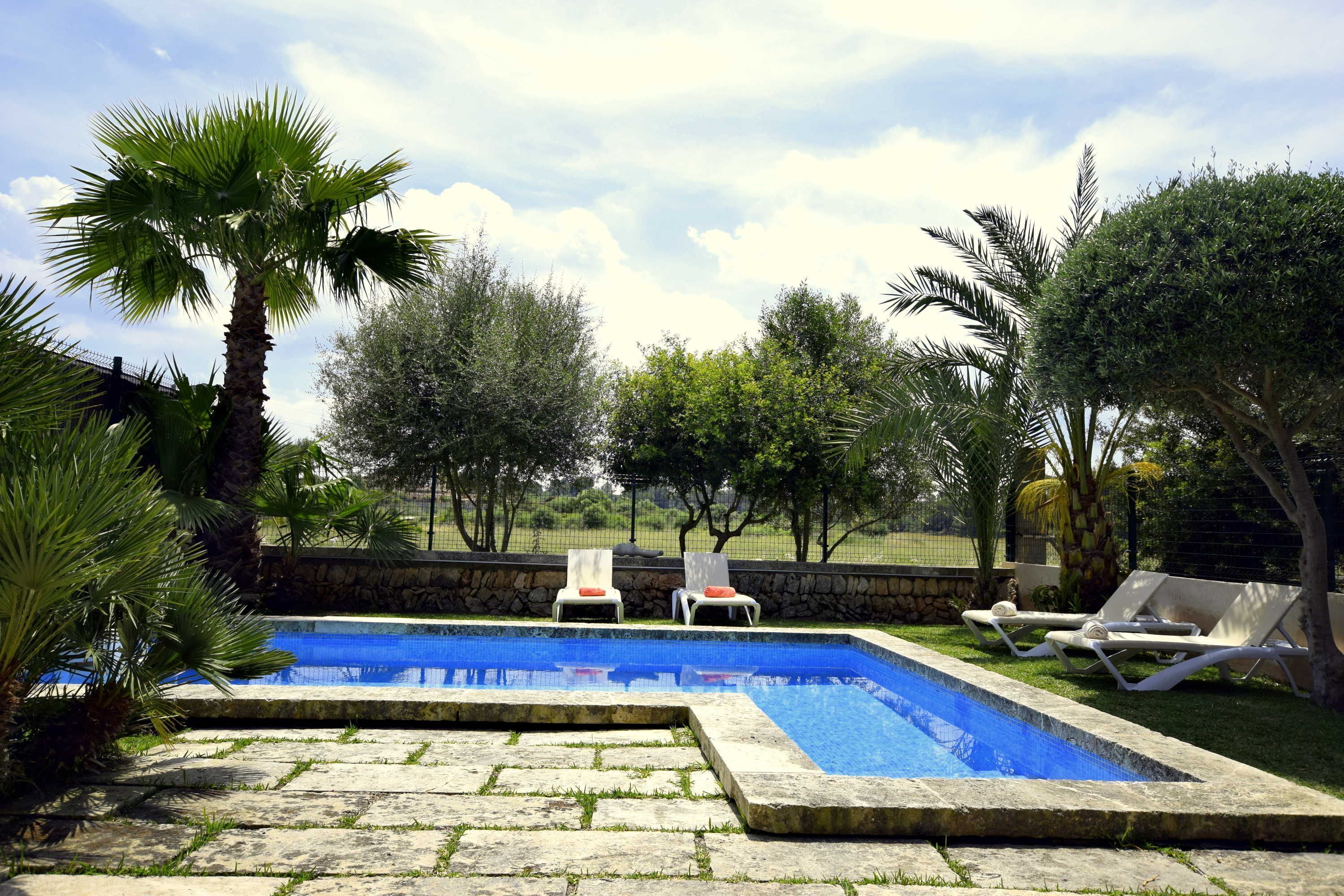 A rectangular swimming pool with a stone border, surrounded by a stone wall and a grassy area. There are two white lounge chairs with orange cushions on the right side of the pool.