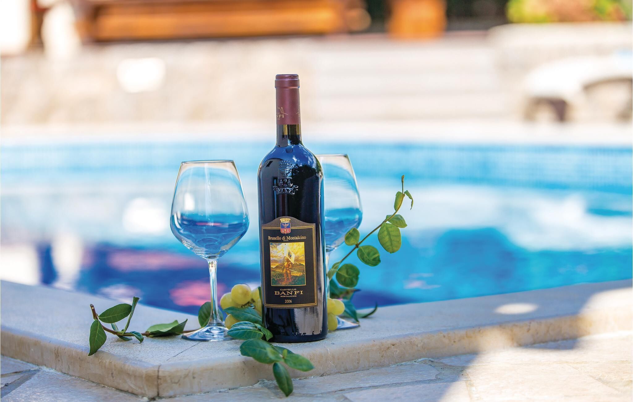 Une bouteille de vin Brunello di Montalcino, deux verres à vin et un décor en bord de piscine agrémenté de verdure.