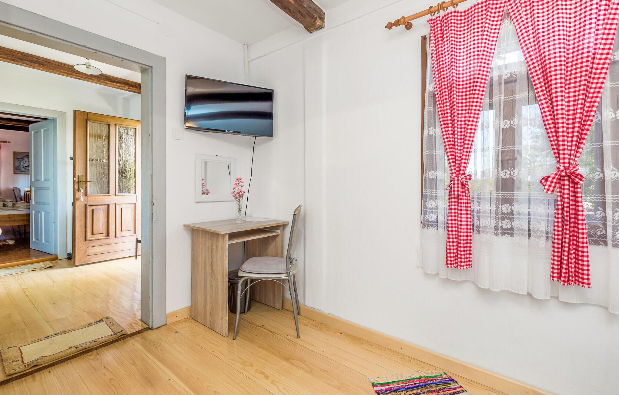 A room with a wooden desk, a chair, a flat-screen TV, and a window with red and white checkered curtains.