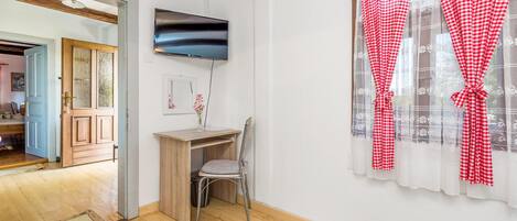 A room with a wooden desk, a chair, a flat-screen TV, and a window with red and white checkered curtains.