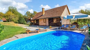 Un'area piscina con lettini e ombrelloni, circondata da una vasca in pietra e una casa in legno con tetto in tegole.