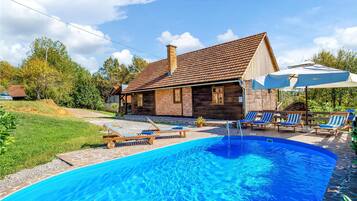 A pool area with lounge chairs and umbrellas, surrounded by a stone pool and a wooden house with a tiled roof.