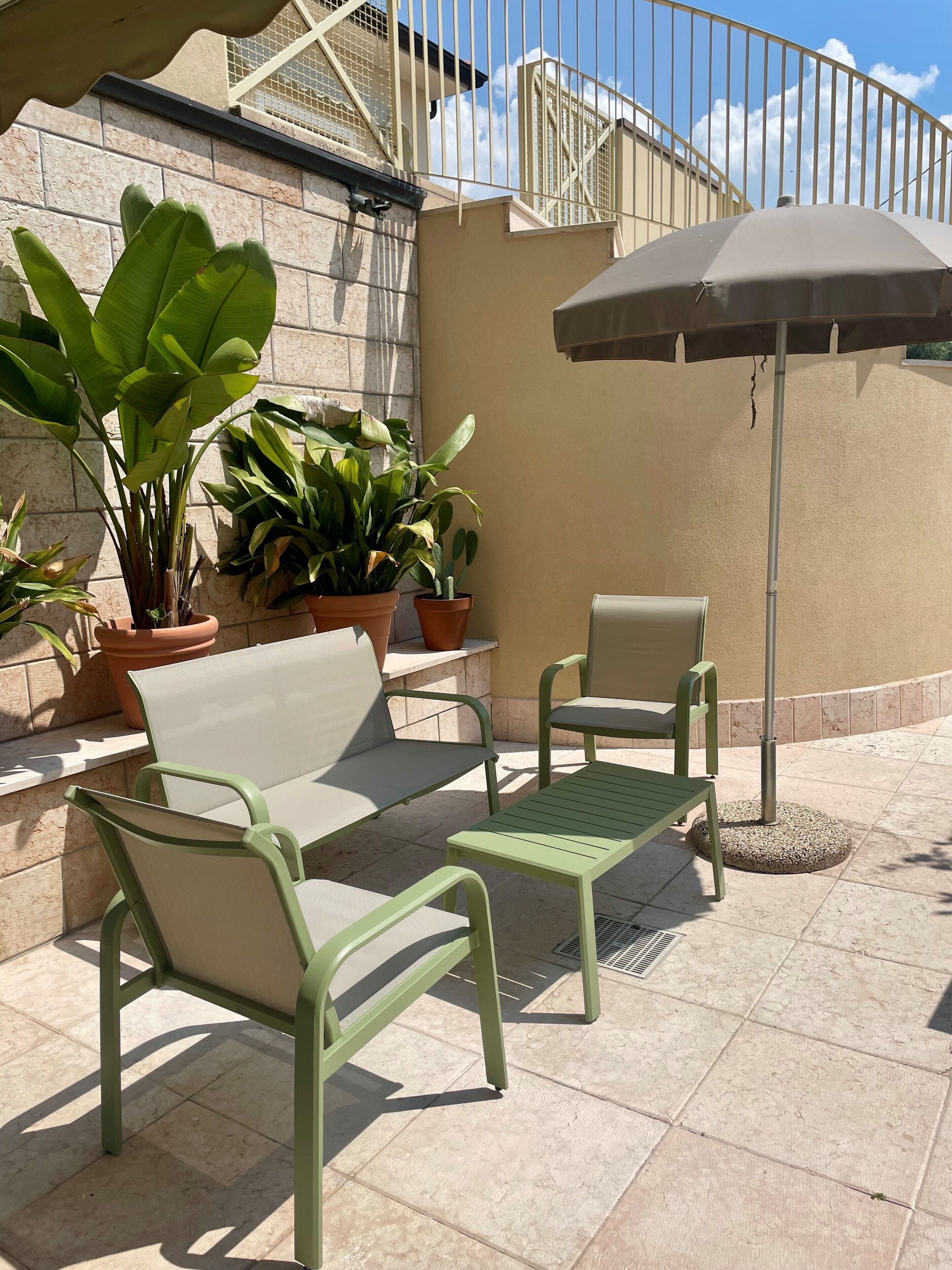 Un patio al aire libre con sillas metálicas verdes y una mesa, rodeado de plantas en macetas.