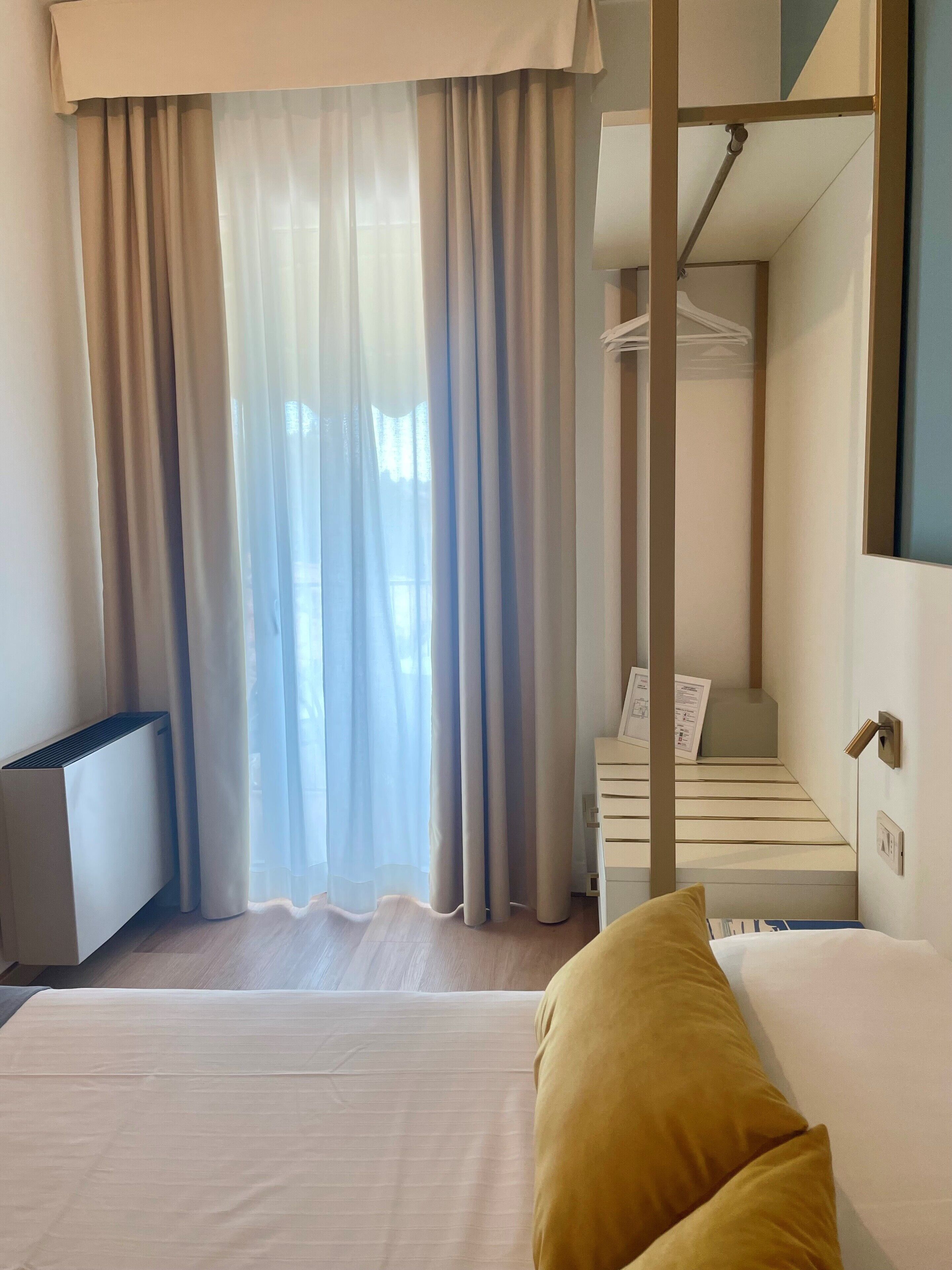 Une chambre d’hôtel comprenant un lit, une table de chevet, un radiateur, une armoire et une fenêtre avec des rideaux.