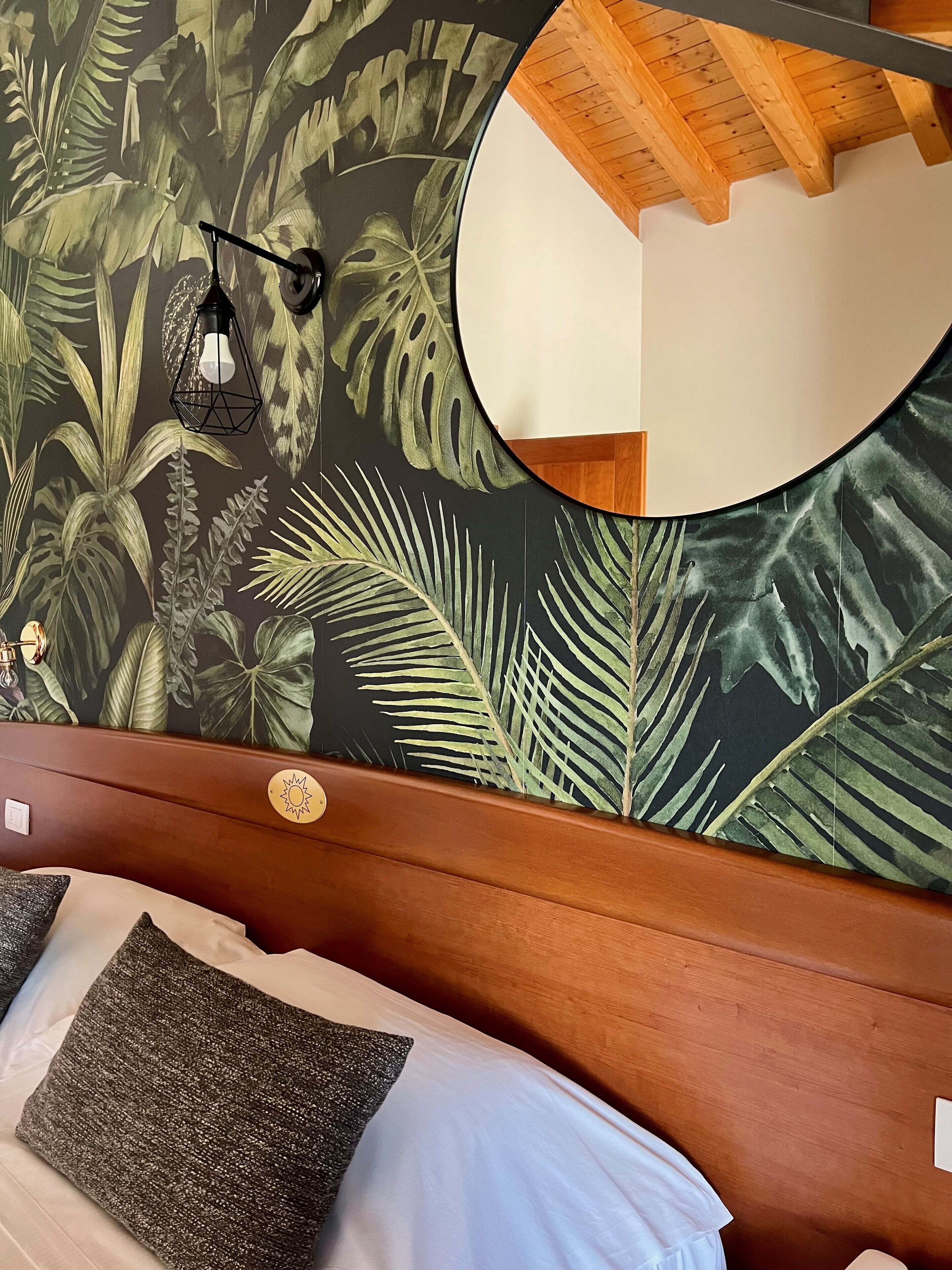 A bedroom with a bed, a wooden headboard, a wall with a tropical leaf pattern, a round mirror, and a hanging light fixture.