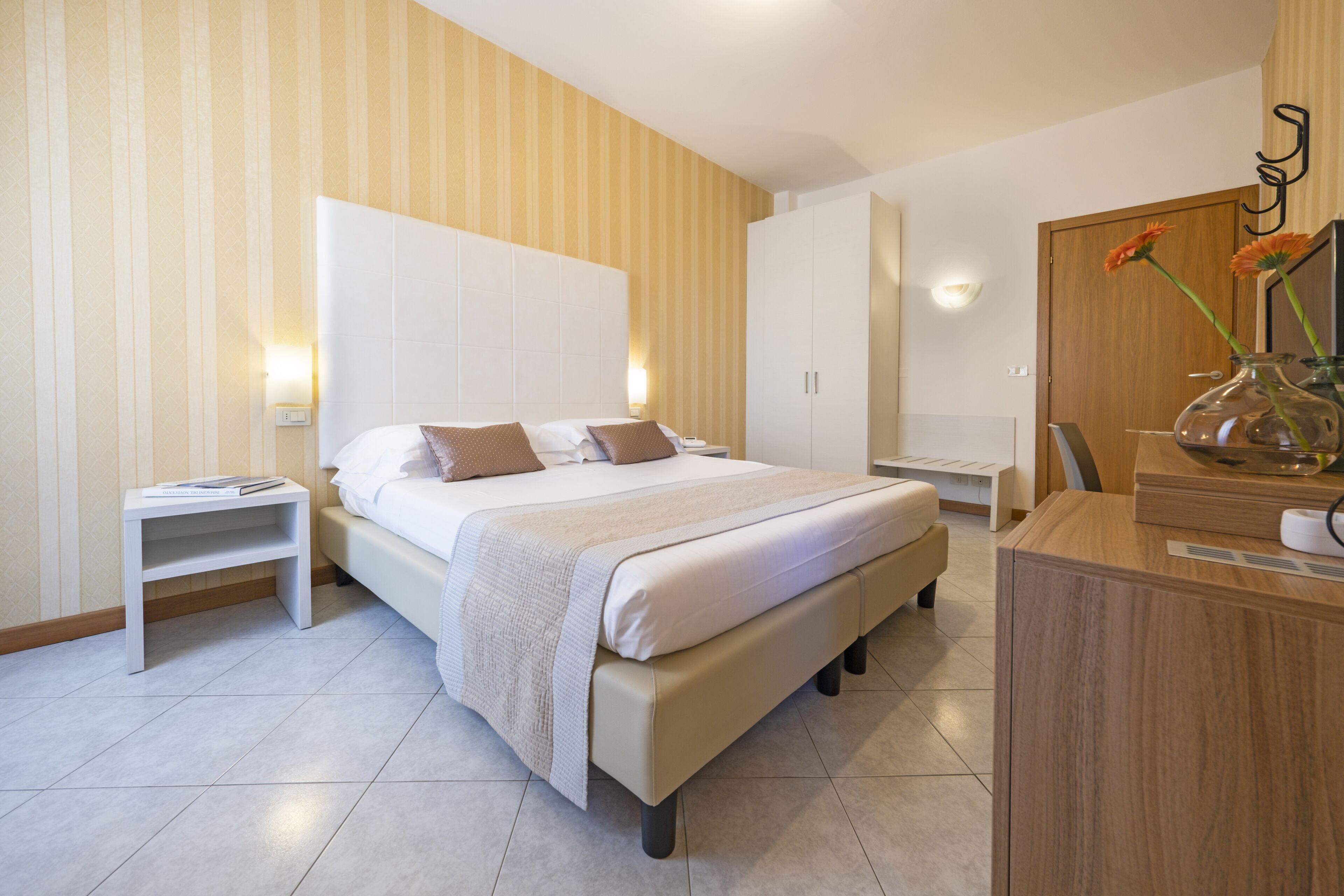 Un dormitorio moderno con una cama grande, mesitas de noche, un armario de madera y un escritorio con un jarrón de flores.