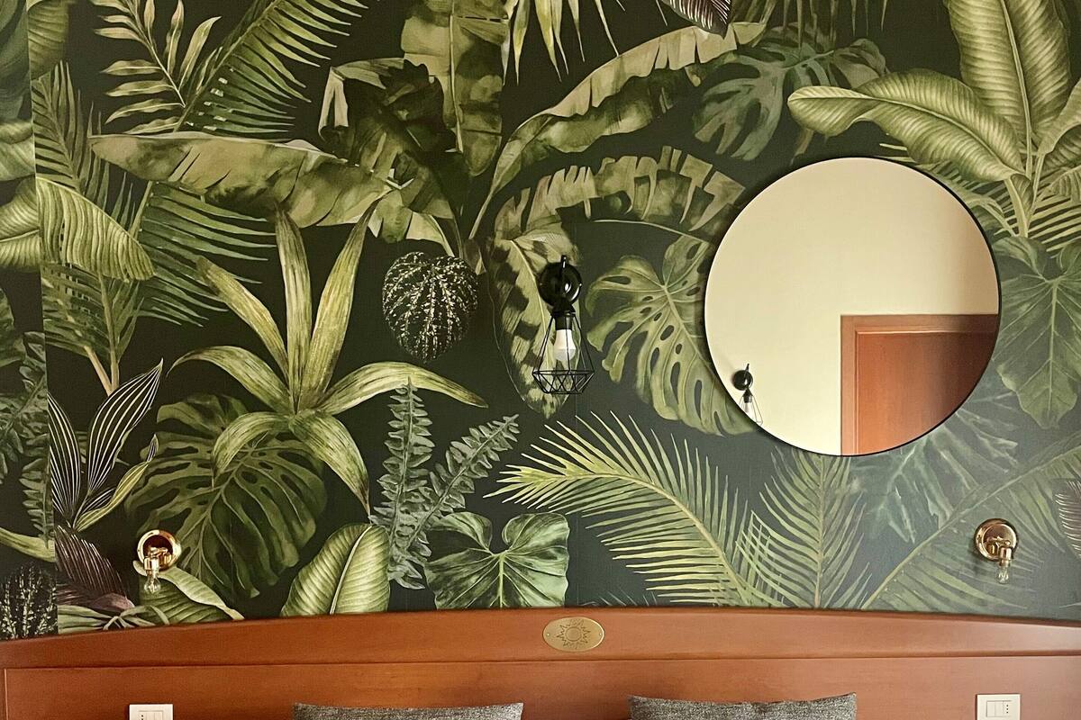 Une chambre d’hôtel avec un lit, des oreillers, un miroir rond et un mur orné d’un motif de feuilles tropicales.