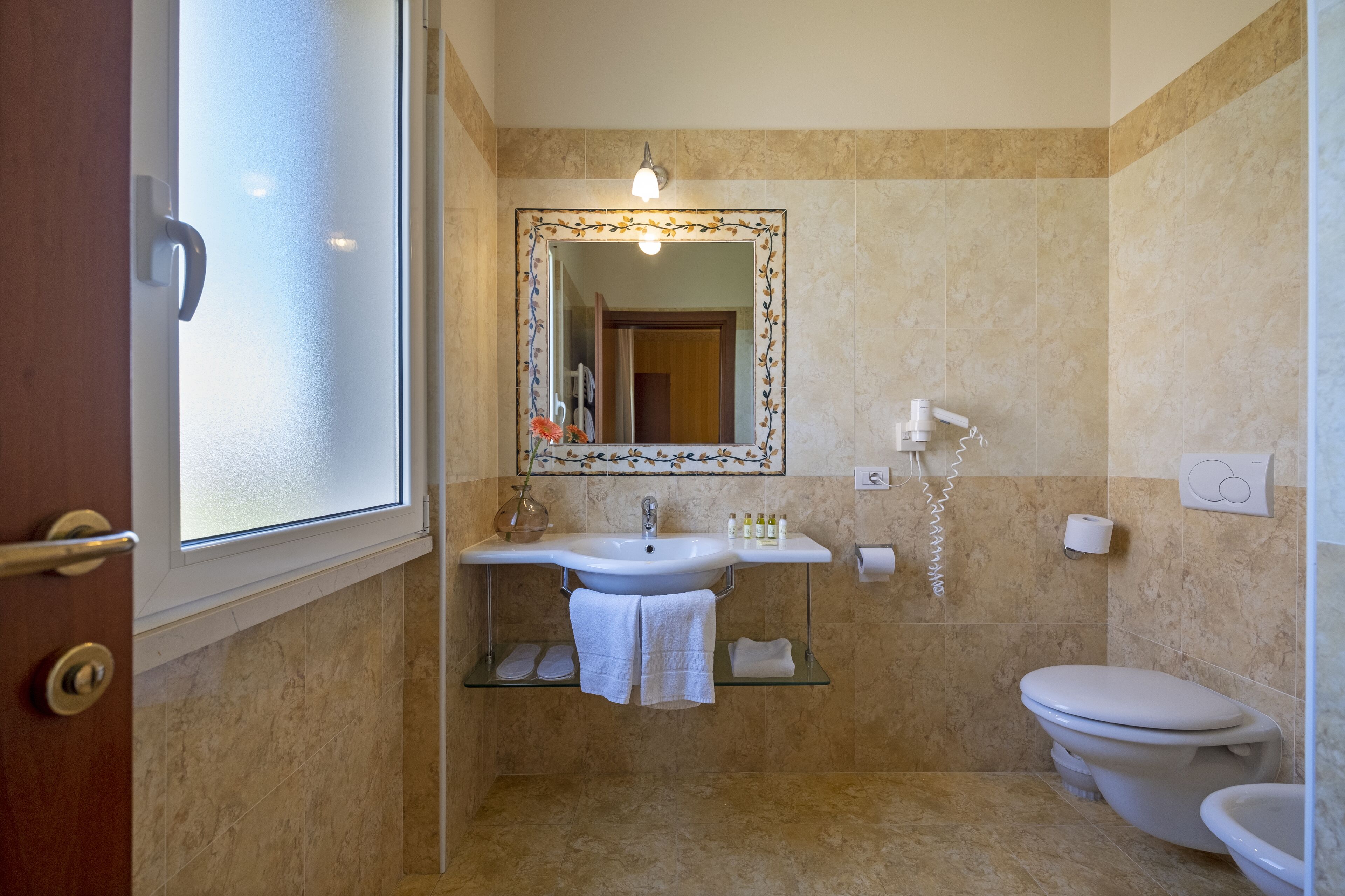 Une salle de bain avec un lavabo blanc, un miroir doté d’un cadre décoratif, des toilettes et une étagère en verre où sont posées des serviettes.
