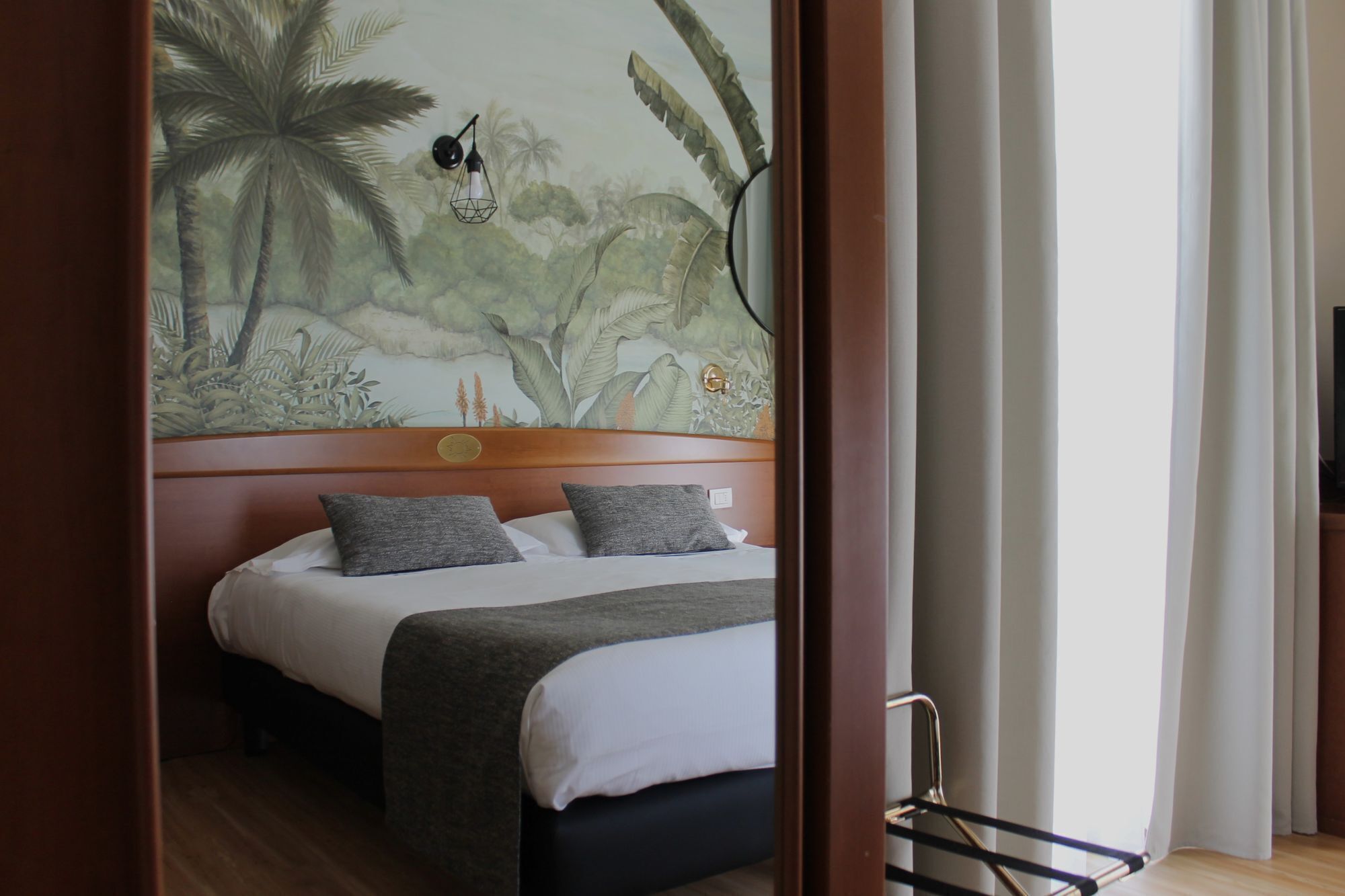 Ein Hotelzimmer mit Bett, einem Kopfteil mit dekorativer Platte, einer Wandmalerei und einem Fenster mit Vorhängen.
