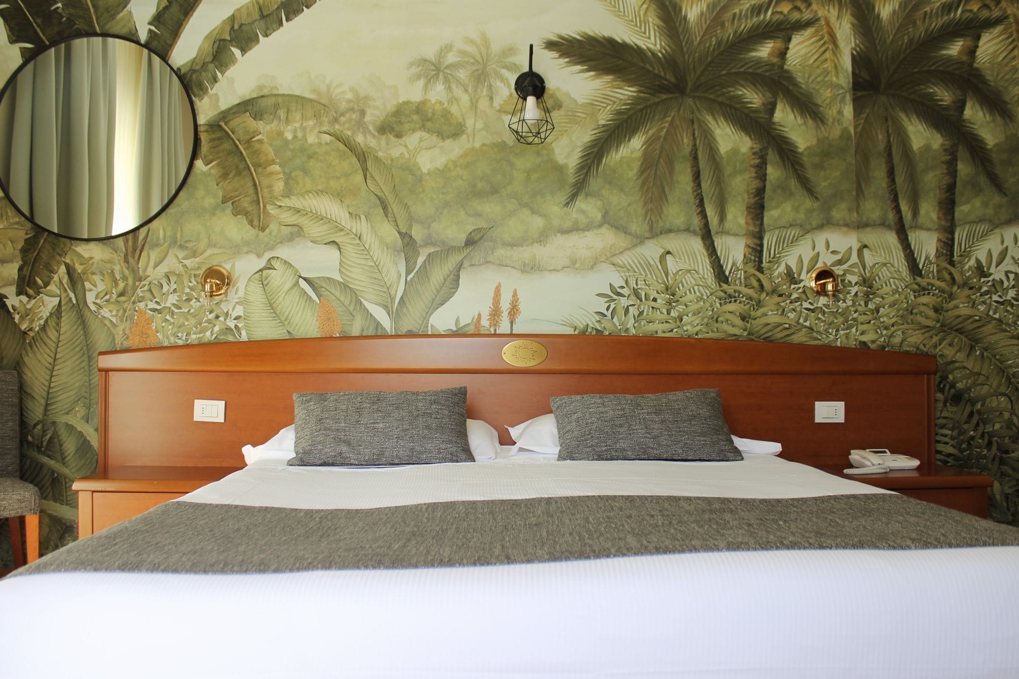 A bed with a wooden headboard, two pillows, a bedside table, and a wall mural of a tropical scene.
