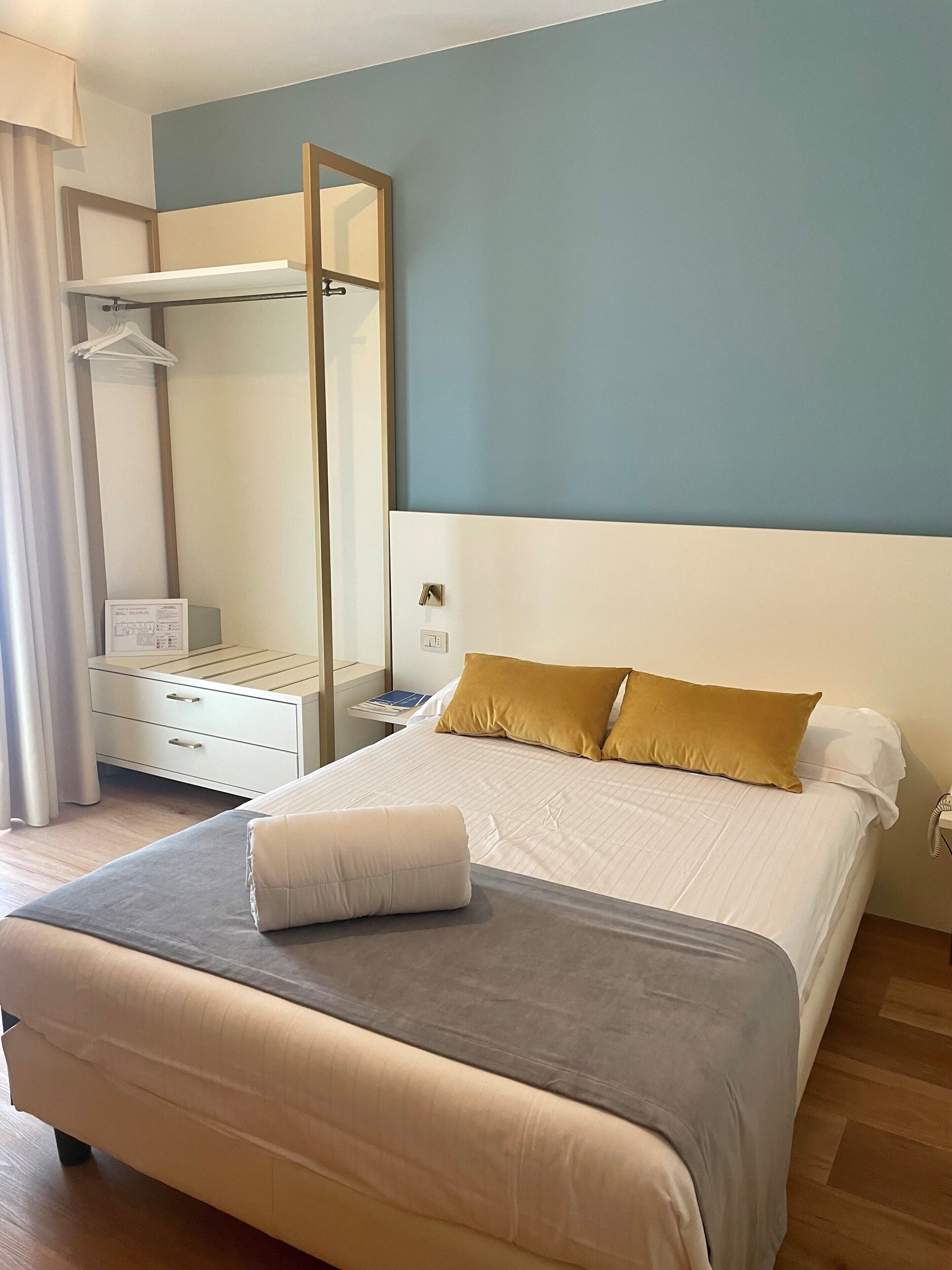 Una habitación de hotel con cama, mesita de noche, espejo, armario y ventana con cortinas.