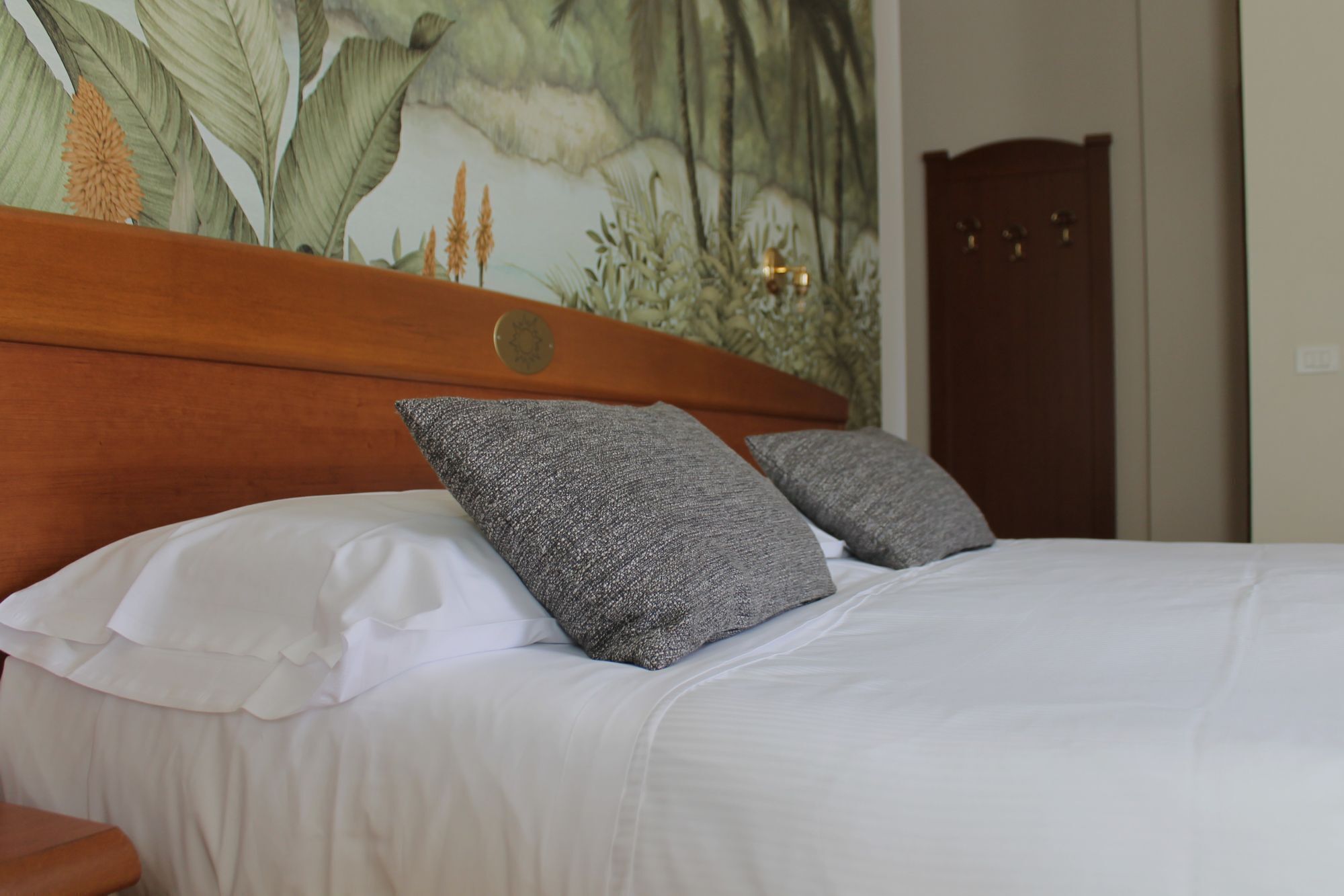 A bed with white bedding and two grey pillows, a wooden headboard, and a wall with a botanical mural.