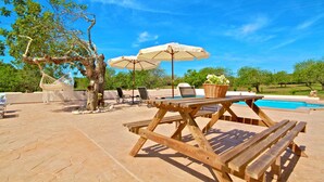 Une terrasse extérieure avec une table et des bancs en bois, entourée de parasols et de chaises, et une piscine en arrière-plan.