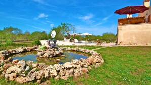 Un jardin avec un étang, un espace aménagé pour s’asseoir à l’extérieur et un bâtiment surmonté d’un parasol rouge.