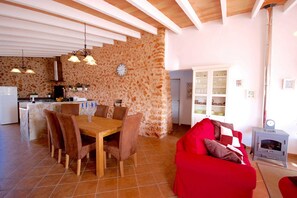 A spacious living area with a red sofa, a wooden dining table with chairs, a kitchen in the background, and a stone wall.