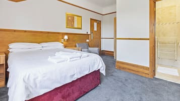 A hotel room with a large bed, a wooden headboard, a grey armchair, a wooden door, and a tiled shower area.