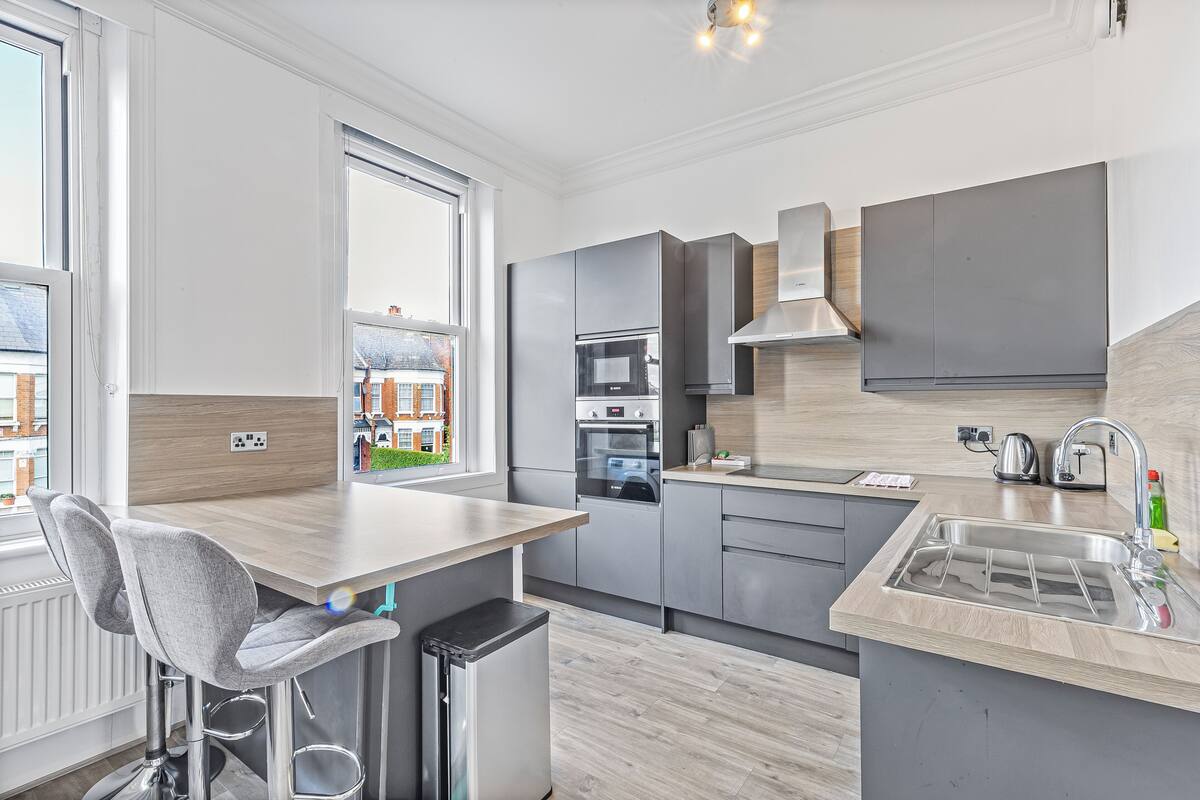A modern kitchen with grey cabinets, a built-in oven, a stainless steel sink, and a window overlooking a cityscape.