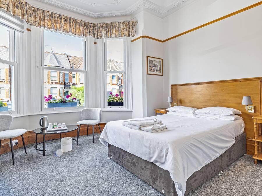 Habitación de hotel con una cama grande, una mesita con hervidor, una silla y una ventana con cortinas.