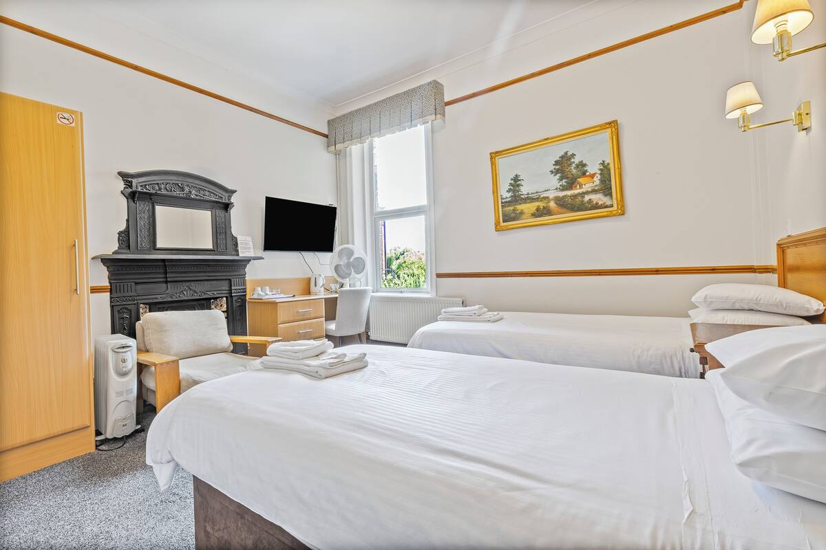 A hotel room with two beds, a desk, a chair, a radiator, a television, and a painting on the wall.