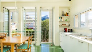 Una cocina moderna con comedor, puertas abiertas que dan a un balcón con vistas a la montaña, y una pequeña isla con fregadero.
