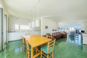 A spacious kitchen with a large wooden table and chairs, a dining area with a bench, and a view of the sea through large windows.