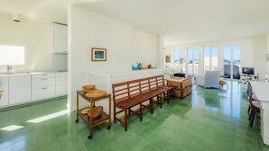 Amplia sala de estar con piso de baldosas verdes, un banco de madera, una mesa de comedor y una cocina con gabinetes blancos y ventanal con vista a la ciudad.