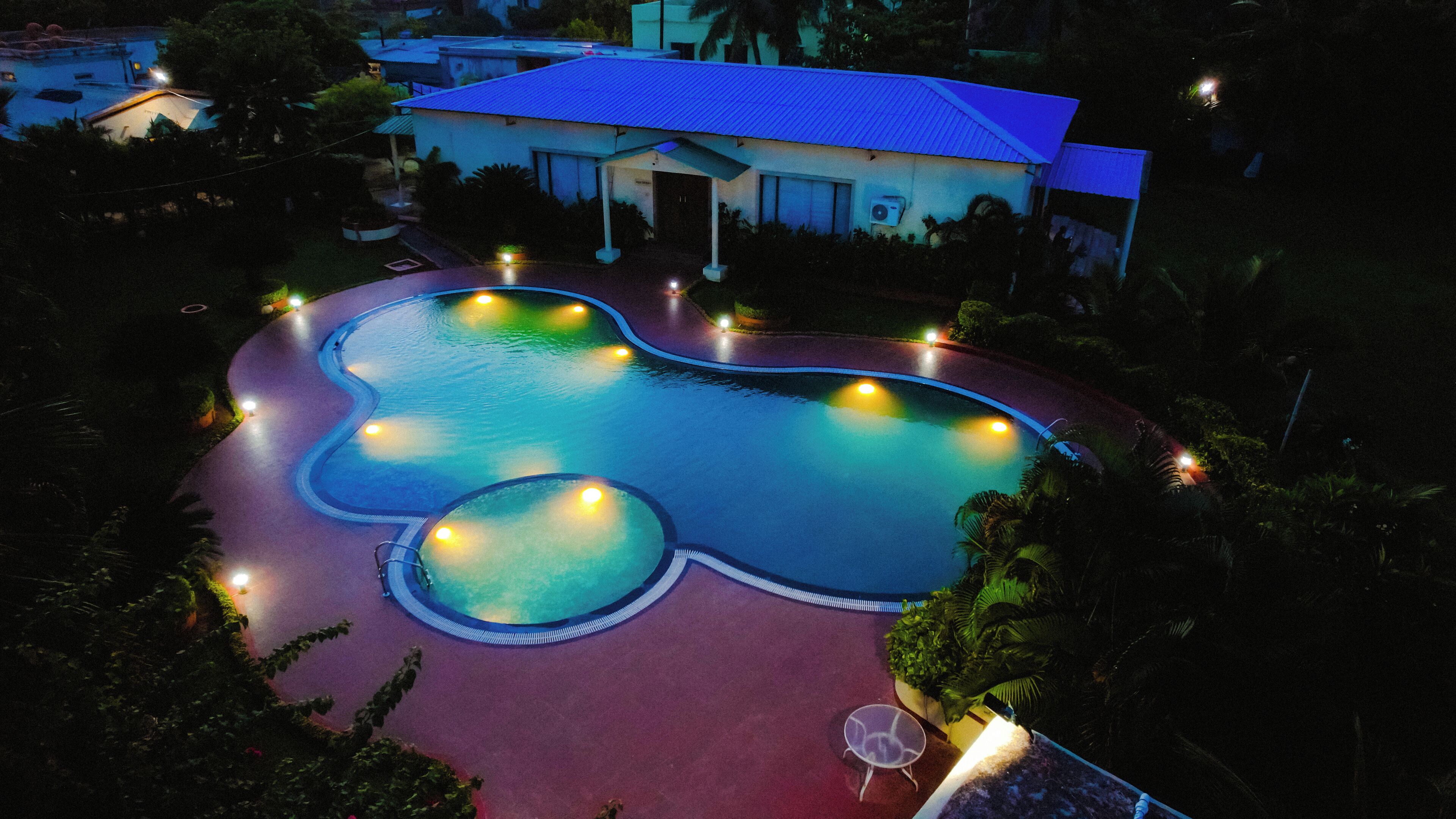 A swimming pool with a unique shape surrounded by a red deck, a house with a blue roof, and a well-lit area.