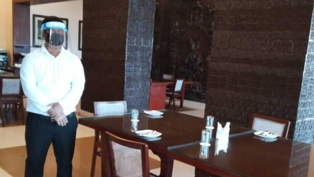 A dining area with a large rectangular table, multiple chairs, and a person standing in the background. - Chanakya BNR hotel (Puri)