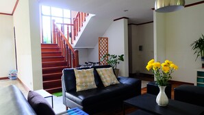 A modern living room with a black leather sofa, a blue ottoman, a black coffee table, and a white vase with yellow flowers. A wooden staircase with red railings leads to an upper level. - Villa Franjipani Maerim (Mae Rim)
