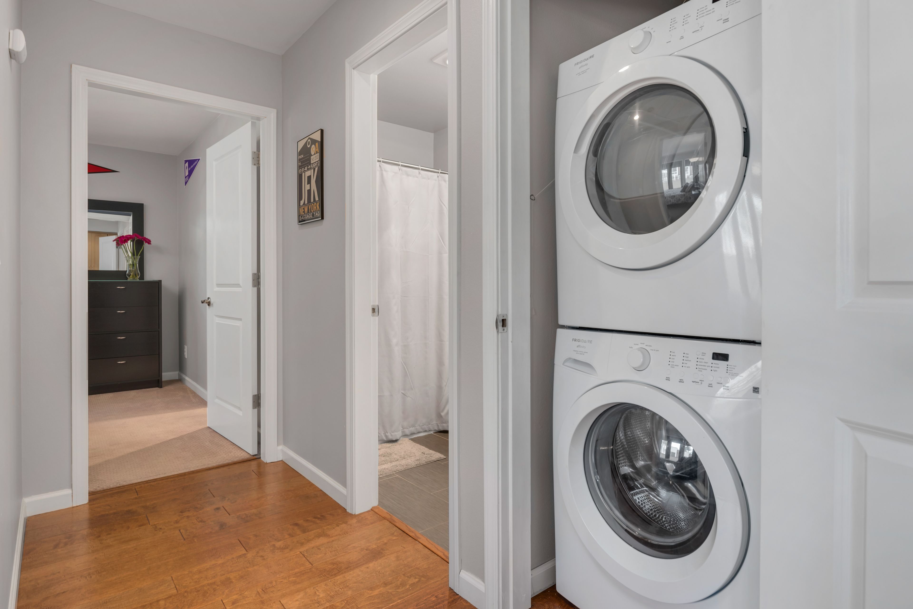 A laundry room with two stacked front-loading washing machines, a wooden floor, and a hallway leading to a bathroom with a shower curtain.