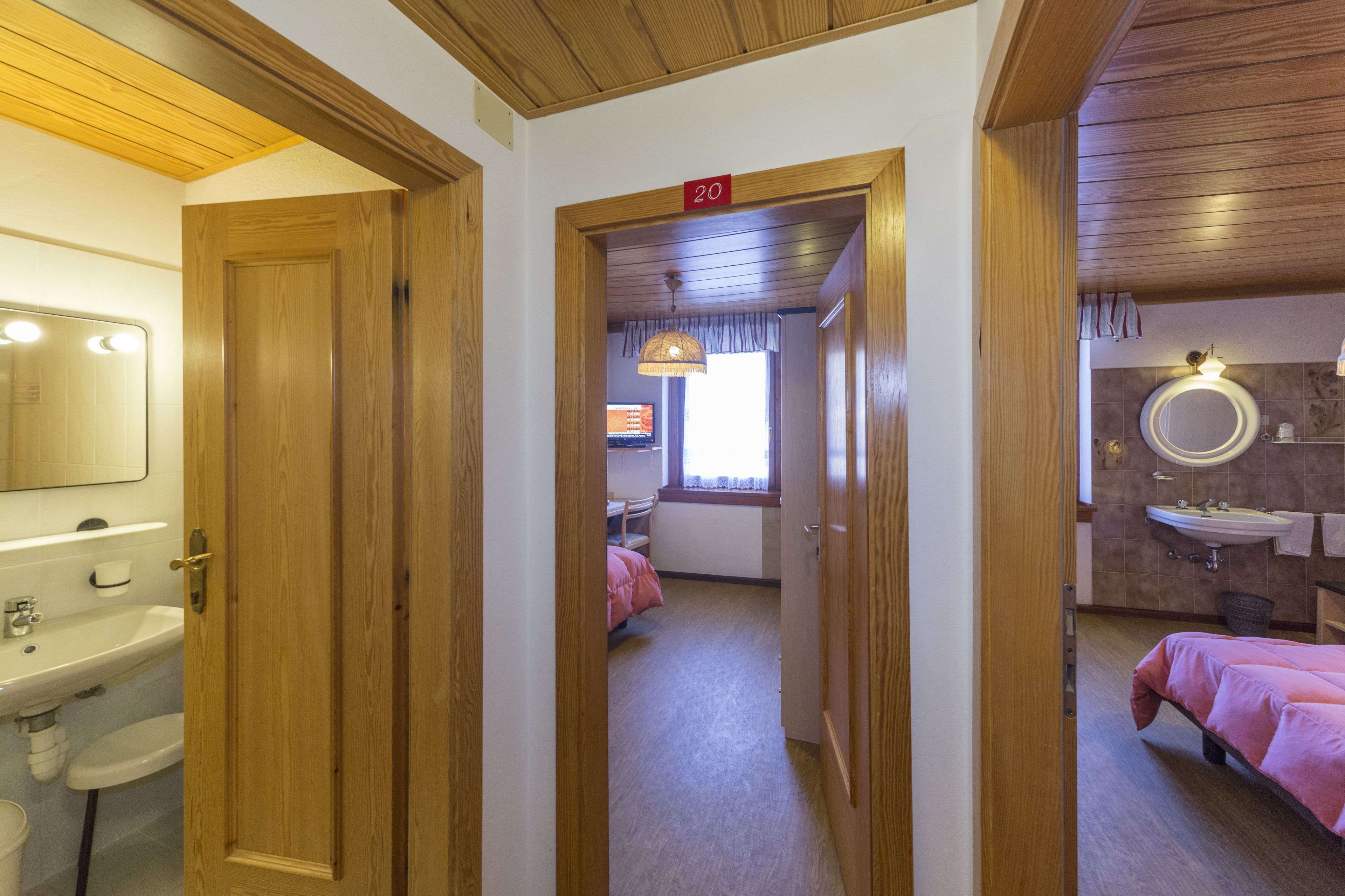 A hotel room with a wooden door, a bathroom with a sink and mirror, a bed with pink bedding, and a number '20' sign on the door.