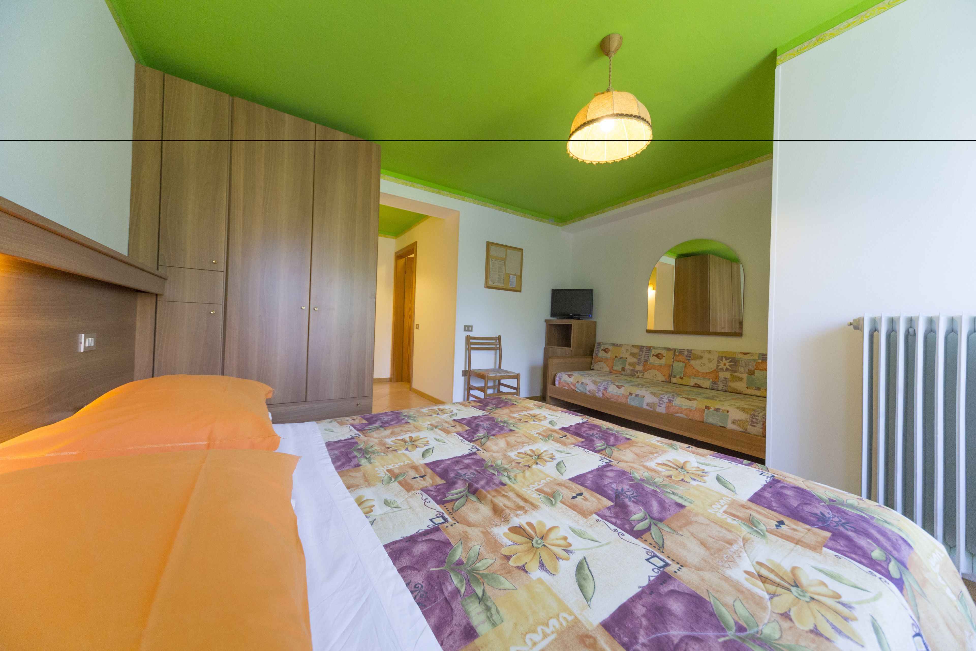 A bedroom with a bed, a wooden wardrobe, a green ceiling, a television, a chair, a sofa, and a radiator.