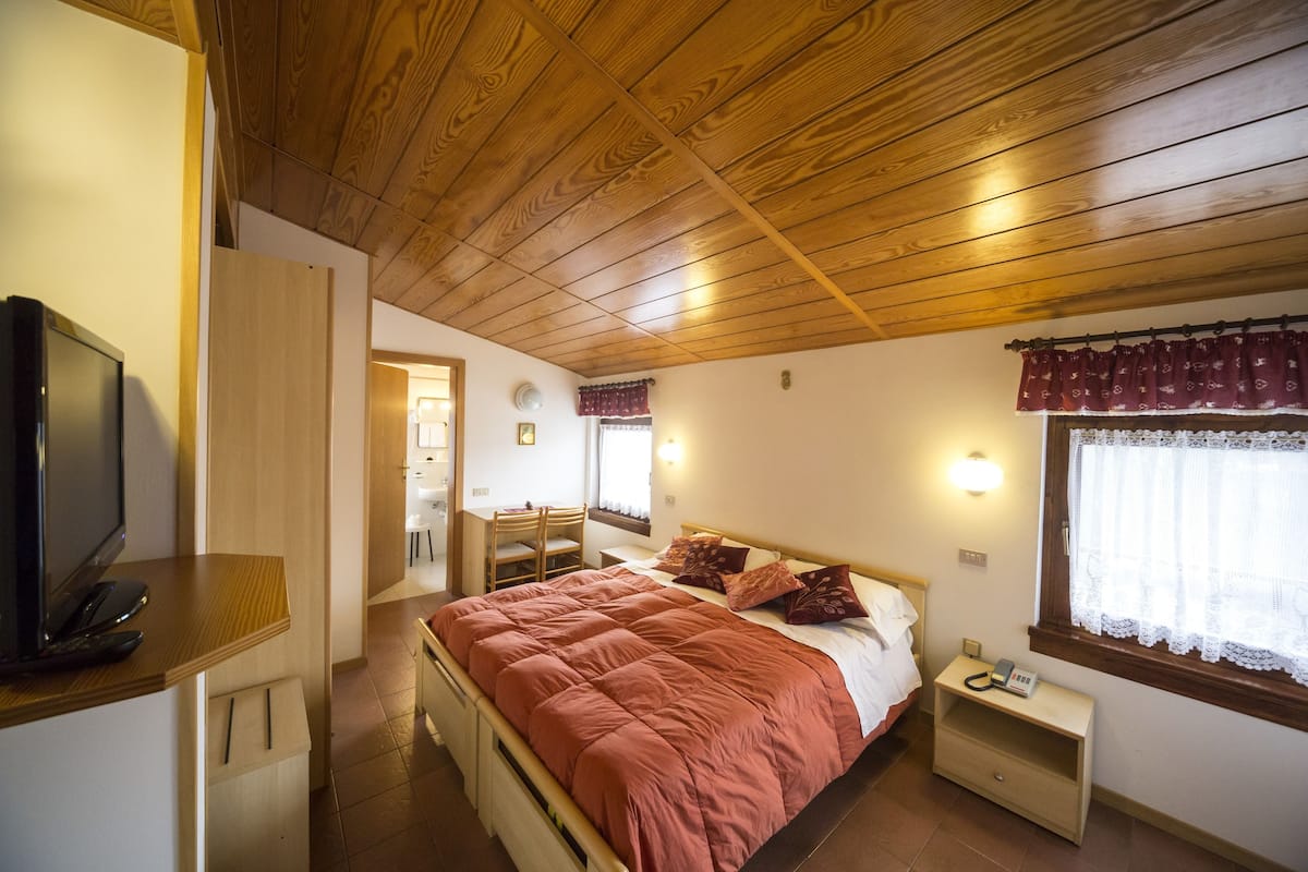 A bedroom with a wooden ceiling, a bed with a red blanket, a bedside table with a lamp, and a TV on a stand.