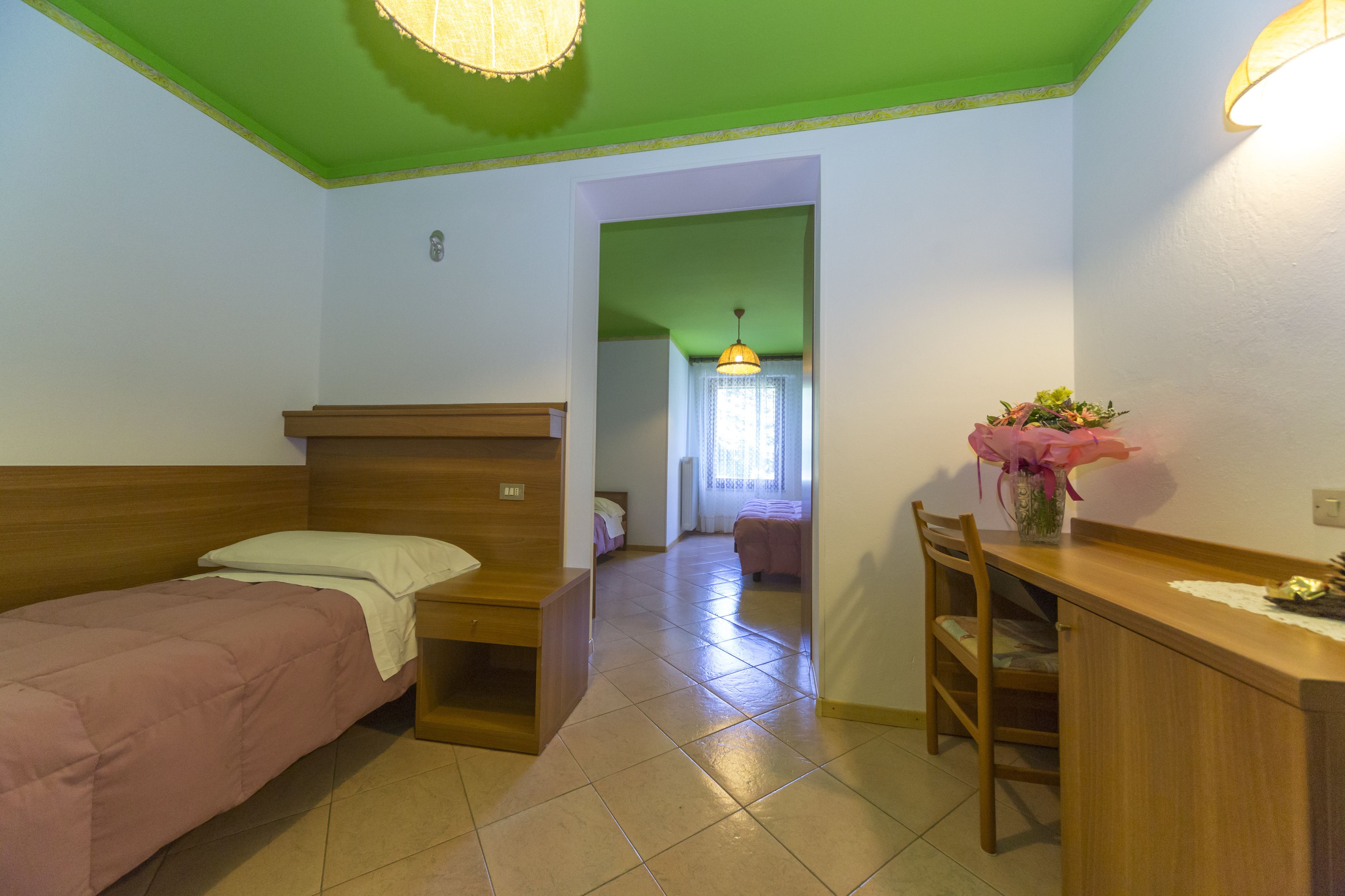 A room with a bed, a desk, and a chair. There is a window with curtains and a hanging light fixture.